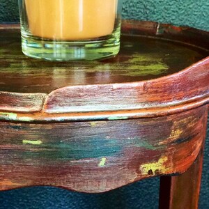 May include: A small, round, wooden side table with a distressed, multi-colored paint finish. The table has a single leg and a curved top. A glass of orange juice sits on the table.