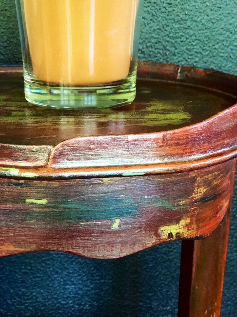 May include: A small, round, wooden side table with a distressed, multi-colored paint finish. The table has a single leg and a curved top. A glass of orange juice sits on the table.