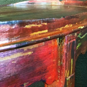 May include: A distressed wooden side table with a red, gold, and green paint finish. The table has a rounded top and a single leg.