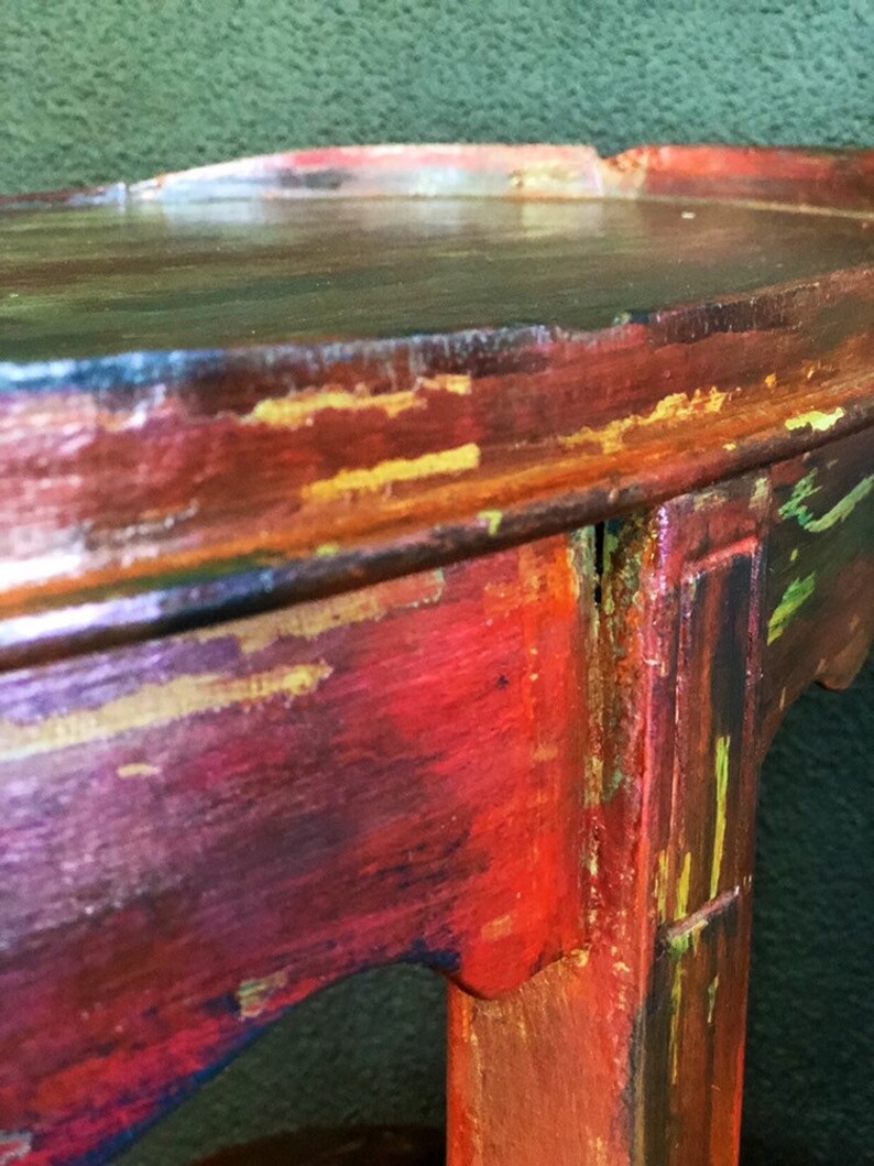 May include: A distressed wooden side table with a red, gold, and green paint finish. The table has a rounded top and a single leg.