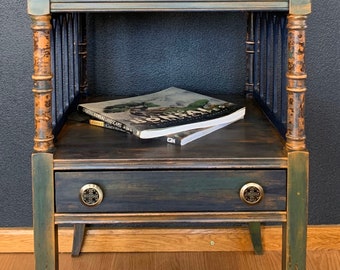 May include: A dark blue and green end table with a distressed finish. The table has a single drawer with brass knobs and a shelf. A book titled "Bonsai Landscapes" is on the shelf.