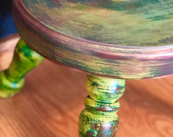 May include: A small, three-legged stool with a round top. The stool is painted in shades of green, yellow, and brown, with a distressed finish. The legs are turned and have a decorative knob at the top.