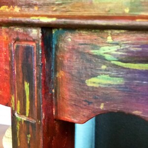 May include: Close-up of a wooden table leg with a multi-colored paint finish. The leg is painted in shades of red, green, yellow, and blue. The paint is applied in a distressed style, giving the table a vintage look.