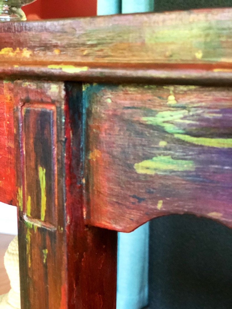 May include: Close-up of a wooden table leg with a multi-colored paint finish. The leg is painted in shades of red, green, yellow, and blue. The paint is applied in a distressed style, giving the table a vintage look.