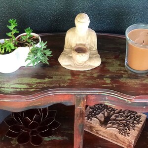May include: A distressed wooden side table with two shelves. The top shelf has a white planter with two succulents, a small Buddha statue, and a brown candle in a glass jar. The bottom shelf has a wooden box with a carved tree design and a metal lotus flower.
