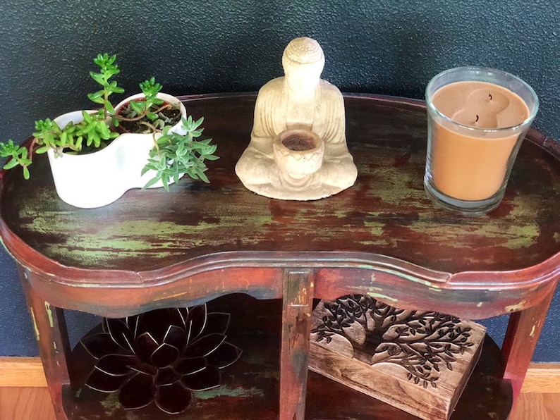 May include: A distressed wooden side table with two shelves. The top shelf has a white planter with two succulents, a small Buddha statue, and a brown candle in a glass jar. The bottom shelf has a wooden box with a carved tree design and a metal lotus flower.