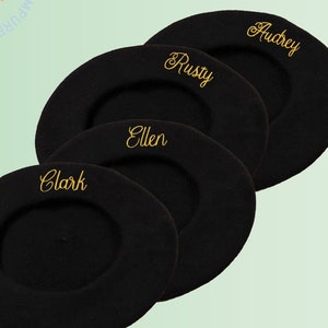 May include: Three black berets arranged in a stack, each featuring gold embroidered cursive text. The names Clark, Ellen, Rusty, and Audrey are stitched onto the fabric. A circular logo with the text Impure Thoughts is visible in the corner.