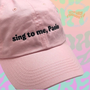 May include: A pink baseball cap with the text "sing to me, Paolo" embroidered in black on the front.