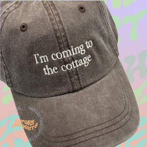 May include: A brown baseball cap with the embroidered text "I'm coming to the cottage" in white. The cap has a curved brim and a classic six-panel design. The hat is made of a soft, worn material.