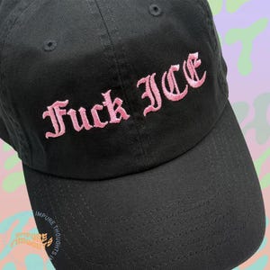 May include: Black baseball cap with the embroidered text "Fuck ICE" in pink gothic lettering. The cap has a curved brim and a structured crown. The phrase is centered on the front of the cap.