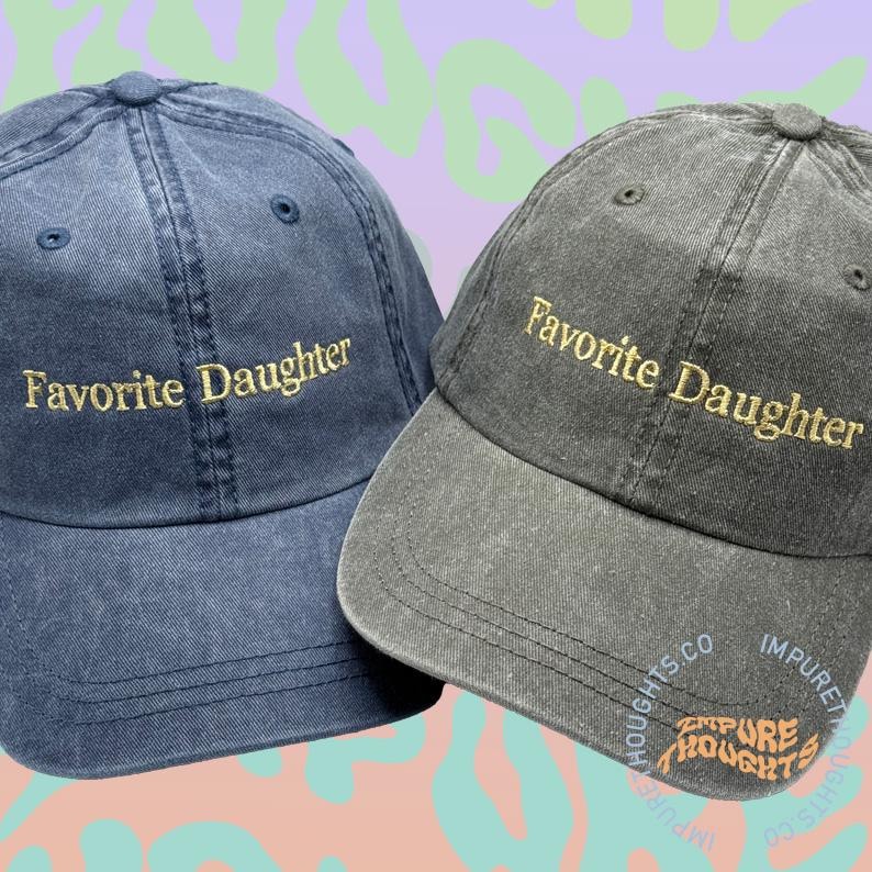 May include: Two baseball caps are shown, one navy blue and one charcoal grey. Both caps feature the embroidered text "Favorite Daughter" in gold thread. The charcoal cap also has the text "Impure Thoughts" in orange.