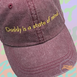 May include: A burgundy baseball cap with the embroidered text "Daddy is a state of mind" in gold thread. The cap has a curved brim and a small button on top. The hat is made of a textured fabric.