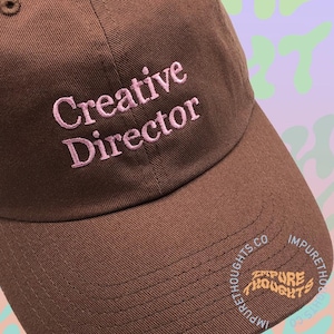 May include: A brown baseball cap with the words "Creative Director" embroidered in pink. The cap has a curved brim and a small logo on the side that says "Impure Thoughts Co."