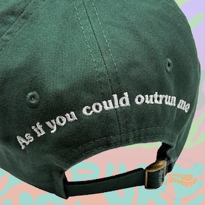 May include: Dark green baseball cap with the embroidered phrase "As if you could outrun me" in white. The cap has a curved brim and an adjustable strap with a brass-colored buckle.
