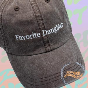 May include: A brown baseball cap with the embroidered text "Favorite Daughter" in white. The cap has a curved brim and a small button on top. The brand name "Impure Thoughts" is printed on the side.