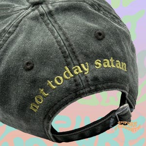 May include: A dark gray baseball cap with the embroidered phrase "not today satan" in gold lettering across the back. The adjustable strap features the text "PURE THOUGHTS" and the brand name "EMPIRE THOUGHTS".