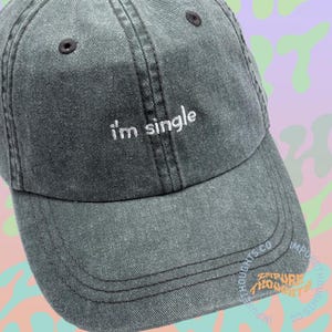 May include: A dark gray baseball cap with the embroidered text "i'm single" in white. The cap has a curved brim and a classic six-panel design. The hat is made of a soft, washed fabric.