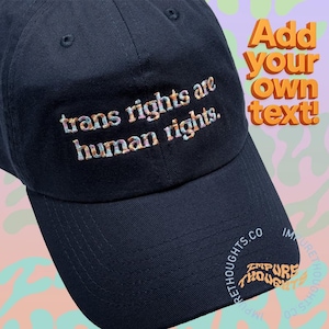 May include: A dark navy blue baseball cap featuring the embroidered text "trans rights are human rights." in a multicolored, variegated thread. The cap includes a curved brim and a structured crown with visible stitching details.
