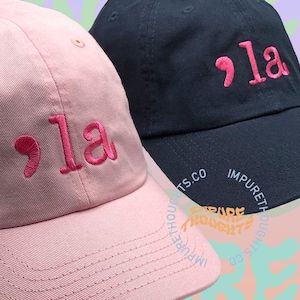 May include: Two baseball caps, one pink and one navy blue, are displayed. Each cap features the embroidered text ",la" in a contrasting pink color. The caps are part of the "Impure Thoughts" collection.