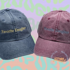 May include: Two baseball caps, one denim blue and one burgundy, embroidered with "Favorite Daughter" in gold and light blue thread. The caps have a classic six-panel design with a curved brim. The background is a mix of purple and green.