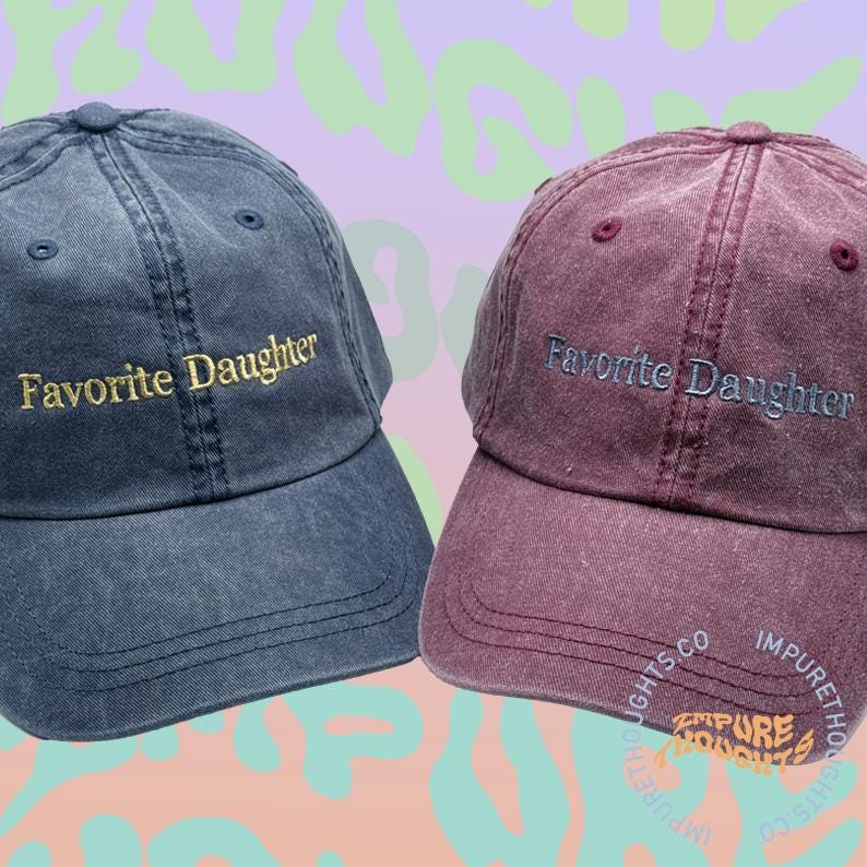 May include: Two baseball caps, one denim blue and one burgundy, embroidered with "Favorite Daughter" in gold and light blue thread. The caps have a classic six-panel design with a curved brim. The background is a mix of purple and green.