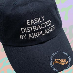 May include: A black baseball cap with white embroidery that reads "EASILY DISTRACTED BY AIRPLANES". The cap has a logo on the back that reads "IMPURE THOUGHTS.CO IMPURE THOUGHTS".
