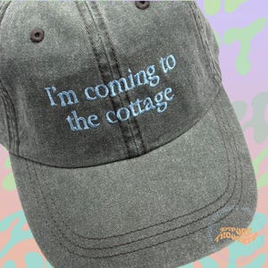 May include: A gray baseball cap with the embroidered text "I'm coming to the cottage" in light blue. The cap has a curved brim and a brown button on top.