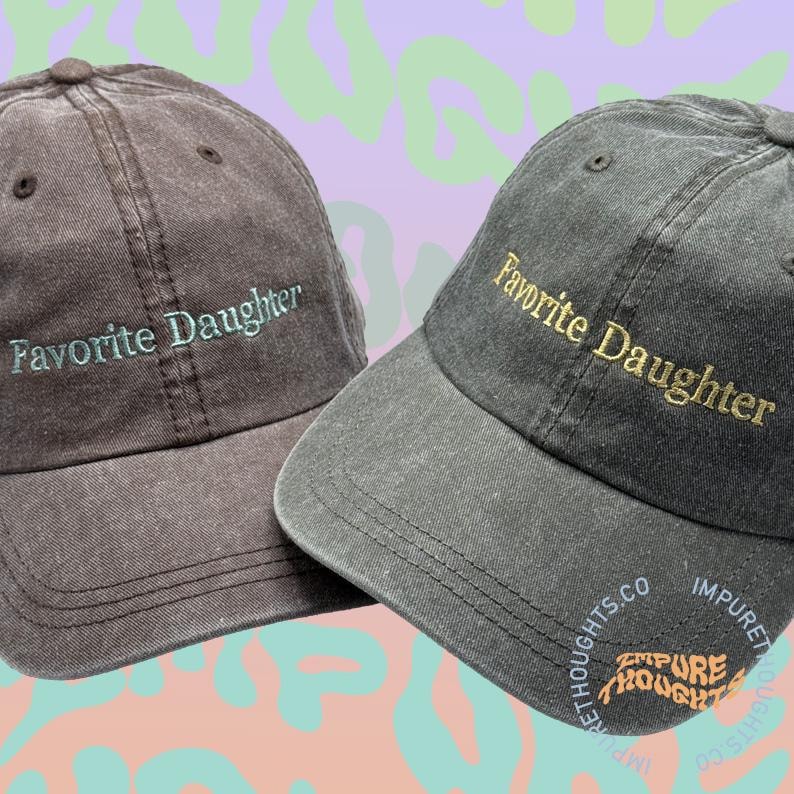 May include: Two baseball caps. One is dark brown with "Favorite Daughter" embroidered in light green. The other is dark grey with gold embroidered text. Both caps have curved brims. The caps have the text "Impure Thoughts" on the side.