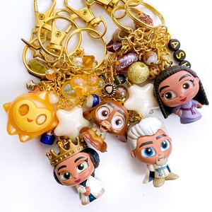 May include: Gold-toned keychains with various charms, including cartoon character figurines, star-shaped beads, and decorative beads. The charms are in shades of orange, brown, white, and purple. The keychains have gold clasps and chains.