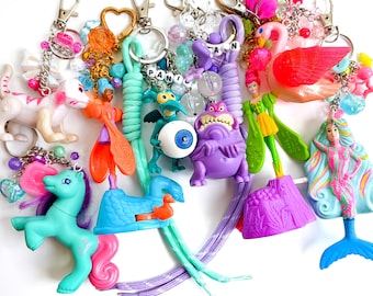 May include: A collection of colorful keychains featuring various plastic figures including a teal pony, a white cat, a purple monster, a mermaid, a flamingo, and a green creature holding an eyeball, all attached to metal rings and lanyards.