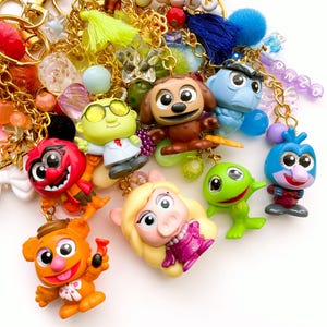 May include: A collection of gold-toned keychains featuring miniature plastic figures of various characters, including a pig, a frog, and a bear. The keychains are adorned with colorful beads, small tassels, and lettered charms spelling out OWL and GONZO.
