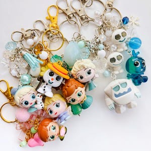 May include: A collection of character keychains featuring various figures from a popular animated film. The keychains have silver and gold-colored clasps, with charms including beads, snowflakes, and character figurines in various colors.