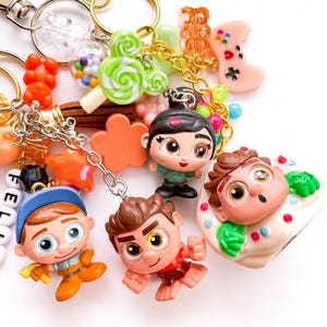 May include: A collection of colorful keychains with various charms. Charms include cartoon character figurines, a green swirled lollipop, a pink video game controller, and orange gummy bears. The keychains have gold and silver metal clasps.