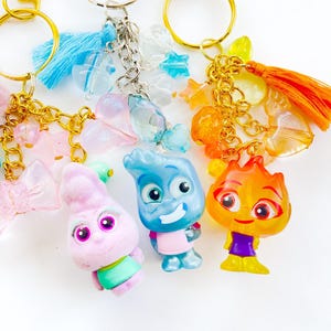 May include: Keychains with character figures and decorative charms. The keychains feature gold and silver-tone metal hardware, colorful tassels, and translucent beads. The character figures are in shades of pink, blue, and orange.