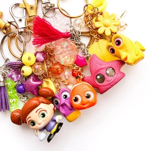 May include: A collection of keychains featuring various colorful plastic charms, including a cartoon girl figure, a clownfish, a pink starfish, a yellow fish, and a purple creature. The charms are attached to gold-tone metal rings with beads, tassels, and lettered cubes spelling FISH.