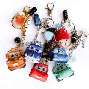May include: Collection of colorful car-shaped keychains. Each keychain features a different colored car, including orange, blue, red, and green. Some keychains have additional decorative elements like beads, charms, and tassels. The keychains are attached to metal clasps.