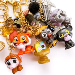 May include: A collection of gold-toned metal keychains featuring various miniature animal figurines, including an orange tiger, a brown monkey, a black panther, and a golden snake, accented with colorful beads, a grey tassel, and a silver feather charm.