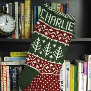 Peut inclure: Une chaussette de Noël avec le nom "CHARLIE" tricoté en blanc. Le bas a un dessus et un talon verts, avec un corps rouge orné de motifs d'arbres et de flocons de neige blancs. Sur une étagère.