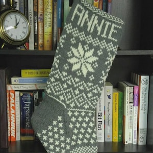 Op de afbeelding: Grijze en witte gebreide kerstsok met de naam "ANNIE" bovenaan. De sok heeft een sneeuwvlokontwerp en andere geometrische patronen. De teen is gebroken wit.