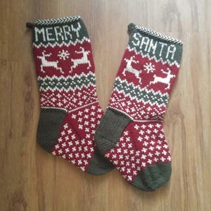 Peut inclure: Deux bas de Noël tricotés. L'un porte l'inscription "MERRY" et l'autre "SANTA" en lettres blanches. Les deux bas ont un fond rouge avec des motifs blancs et verts, dont des rennes et des flocons de neige. Le bout et le revers sont verts.