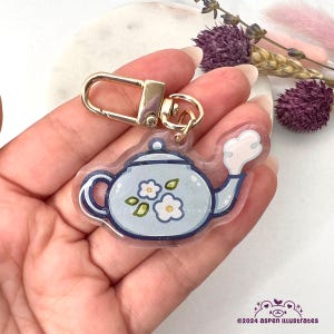 Pode incluir: Um chaveiro de chaleira azul com flores brancas e folhas verdes. A chaleira tem uma alça e um bico dourados. O chaveiro é feito de acrílico transparente e tem um fecho de metal dourado.