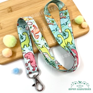 Puede incluir: Un cordón azul claro con un patrón repetido de dinosaurios de dibujos animados coloridos, huevos y follaje. El cordón tiene un cierre de metal plateado y está sobre una superficie de madera. El cordón mide aproximadamente 43 cm de largo.