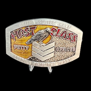 May include: A silver belt buckle with a black crow perched on a yellow and brown wooden plank. The buckle reads "United States Navy First Class Petty Officer" and "Leading From The Front".