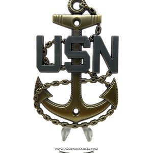 May include: A gold-colored anchor with a chain wrapped around it. The anchor has a gray "USN" in the center. The anchor is on a white background.