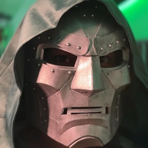 May include: A metallic silver-colored face mask featuring a rigid, angular design with a prominent brow, a sharp nose bridge, and a horizontal mouth slit. The mask is adorned with small, evenly spaced rivets and is paired with a matching fabric hood.