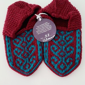 May include: Pair of maroon knitted slippers with a teal geometric pattern. The slippers have a tag attached that reads "Your purchase helps women in rural areas earn income while providing deeper purpose in their lives and preserving an important part of Azerbaijani culture. Size 38 US Women"