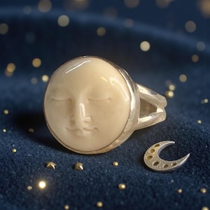 Puede incluir: Un anillo de plata que presenta un rostro de luna circular de color crema con los ojos cerrados y una expresión suave. La banda es de plata pulida. Junto al anillo hay un pequeño dije de media luna plateado con recortes circulares.