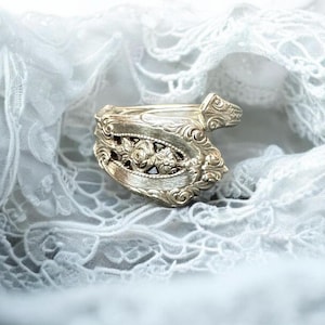 May include: Silver-toned ring with intricate floral and scrollwork details. The ring features a central floral design and ornate embellishments. The ring is set against a backdrop of white lace.
