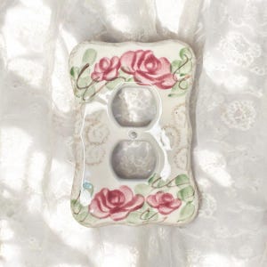 Pode incluir: Uma placa de cerâmica para interruptor de luz com um design floral. A placa é retangular com bordas arredondadas e apresenta dois orifícios para tomadas. Rosas cor-de-rosa e folhas verdes são pintadas sobre um fundo creme.