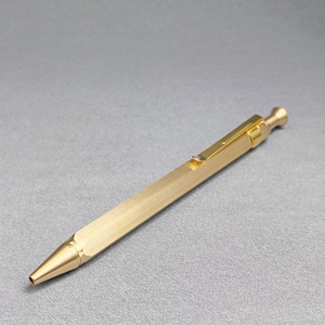 May include: A hexagonal brass mechanical pencil with a metallic gold finish. The writing instrument features a pointed tip, a slim body with a brushed texture, and a matching gold-colored pocket clip attached near the top click mechanism.